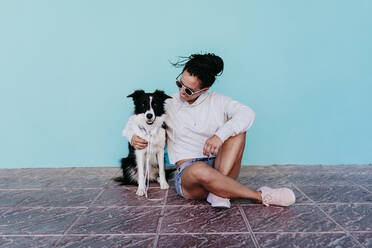 Happy woman sitting with arm around Border Collie dog against turquoise wall - EBBF00824