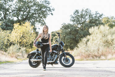 Beautiful biker woman walking while looking away against motorcycle on road - DAMF00516