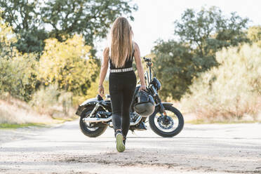 Blond woman holding helmet while walking towards motorcycle on road - DAMF00517