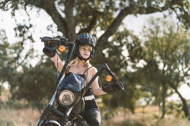 Beautiful woman wearing crash helmet while sitting on motorcycle against tree - DAMF00518