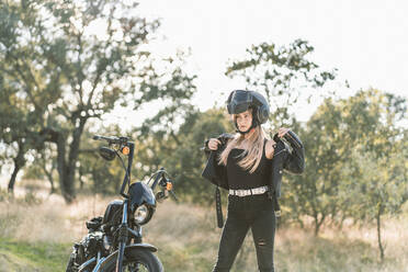 Blond young female biker wearing leather jacket while standing by motorcycle - DAMF00519