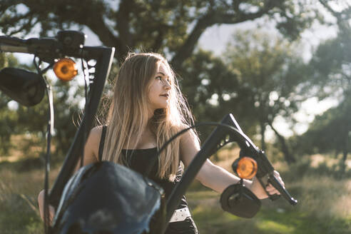 Beautiful blond woman looking away while sitting on motorcycle against tree - DAMF00520