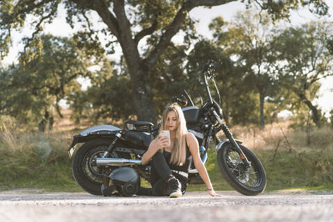 Blond female biker using smart phone while sitting against motorcycle on roadside stock photo