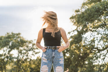 Young woman with long blond tousled hair standing against sky - DAMF00524