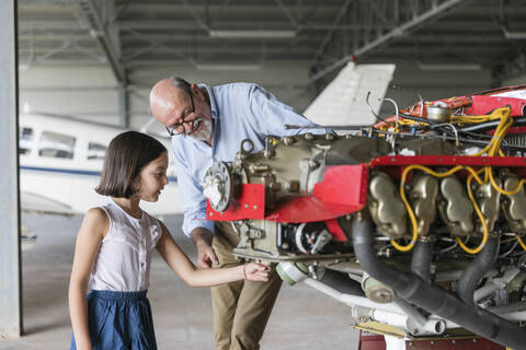 Großvater zeigt seiner Enkelin im Hangar, wie man ein Flugzeug repariert, lizenzfreies Stockfoto