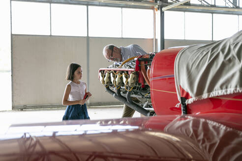 Großvater zeigt seiner Enkelin konzentriert, wie man ein Flugzeug repariert, während er im Hangar steht - EIF00196