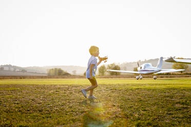 Cute little boy playing with toy plane while standing at airfield on sunny day - EIF00227
