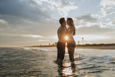 Man an woman kissing while standing in water at beach during sunset - GMLF00665