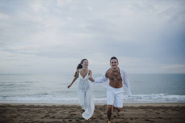 Glückliches Paar hält sich beim Laufen am Strand an den Händen - GMLF00673