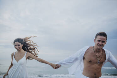 Smiling couple holding hands while running against sea - GMLF00674