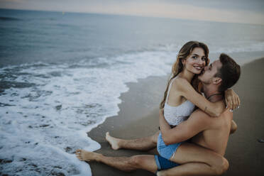 Man kissing woman sitting on his lap at beach - GMLF00694