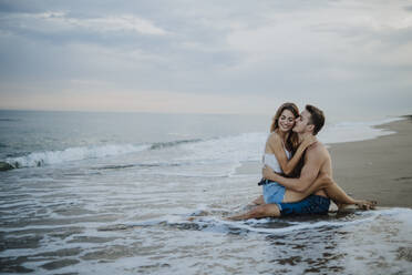 Young man kissing woman sitting on his lap at beach - GMLF00695