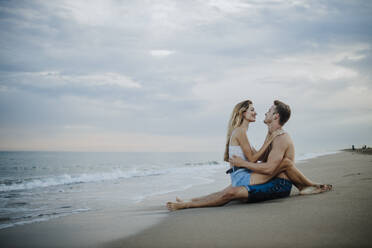 Young couple looking at each other while sitting on beach - GMLF00696