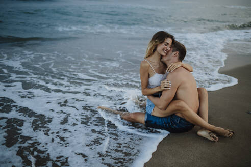 Paar umarmt sich, während es am Strand am Rande des Wassers sitzt - GMLF00697