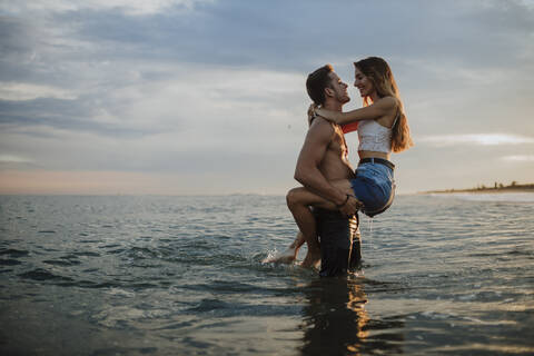 Man carrying woman while standing in water during sunset at beach stock photo
