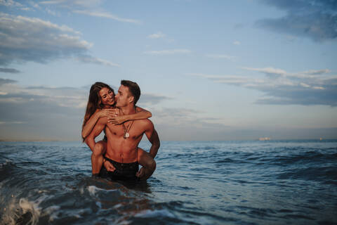 Smiling man piggybacking woman while standing in water at beach stock photo