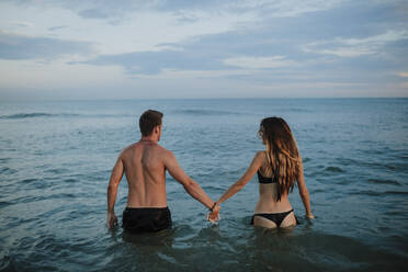 Couple wearing swimwear holding hand while standing in water at beach - GMLF00710