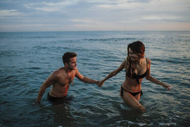 Paar hält sich an der Hand, während es bei Sonnenuntergang am Strand im Wasser spazieren geht - GMLF00712