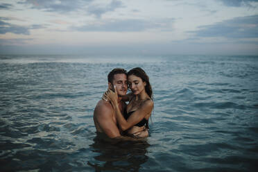 Paar mit geschlossenen Augen im Wasser stehend am Strand bei Sonnenuntergang - GMLF00720