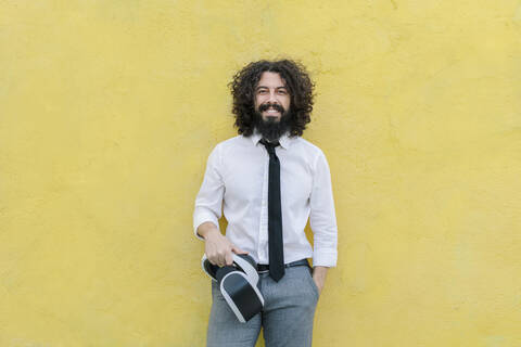 Smiling businessman with hand in pocket holding virtual reality eyeglasses while standing against wall stock photo