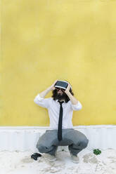 Man wearing virtual reality eyeglasses crouching against wall - MRRF00563