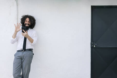 Businessman waving hand to video call on phone while standing against wall stock photo