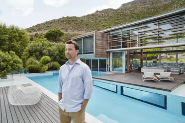 Mature man with hands in pockets looking away while standing against modern house - RORF02339