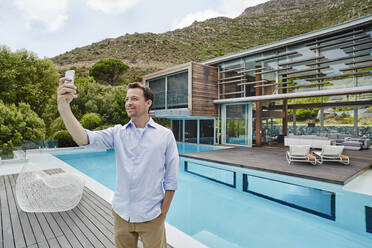 Mature man taking selfie on smart phone while standing against modern house - RORF02342