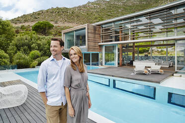 Smiling couple looking away while standing against modern house - RORF02355