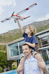 Son sitting on father's shoulder with toy airplane against modern house - RORF02384