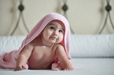 Baby girl in towel looking up while lying on bed at home - KIJF03348