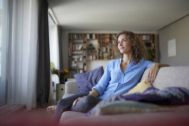 Thoughtful woman smiling while sitting on sofa at home - RBF07943