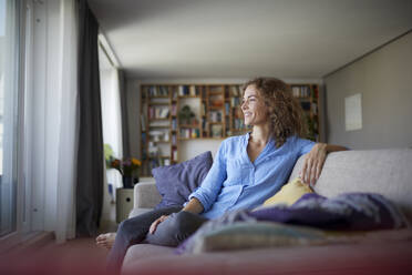 Smiling woman looking away while sitting on sofa at home - RBF07944