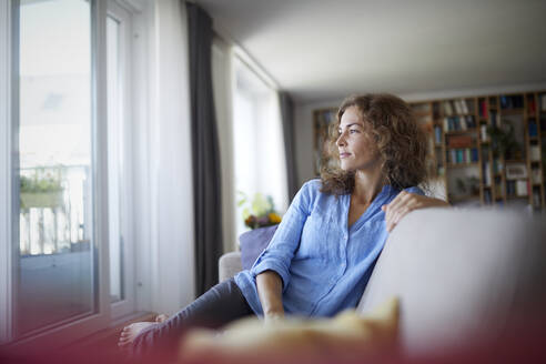 Frau schaut zum Fenster, während sie zu Hause auf dem Sofa sitzt - RBF07945
