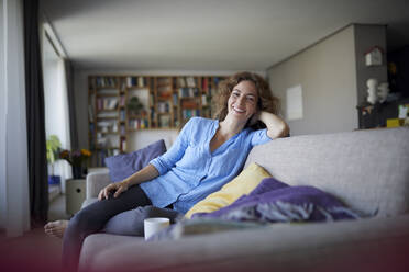 Smiling woman with head in hands sitting on sofa at home - RBF07947