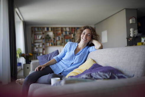 Lächelnde Frau mit dem Kopf in den Händen sitzt zu Hause auf dem Sofa - RBF07947