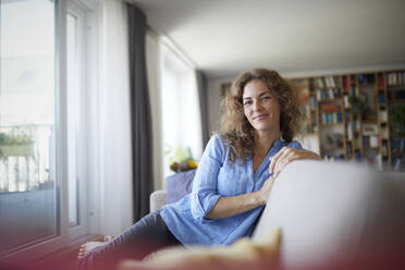 Smiling woman sitting on sofa by window at home - RBF07948