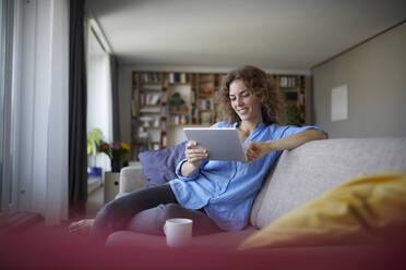 Frau benutzt digitales Tablet, während sie zu Hause auf dem Sofa sitzt - RBF07949