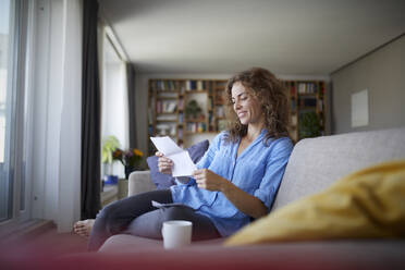 Woman reading paper while sitting on sofa at home - RBF07950