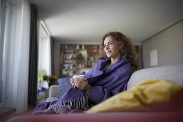 Smiling woman wearing shawl drinking coffee while sitting on sofa at home - RBF07951