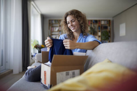 Smiling woman removing cloth from package while sitting on sofa at home stock photo