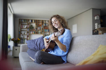 Smiling woman playing guitar while sitting on sofa at home - RBF07954