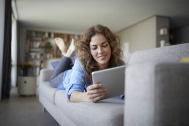 Frau benutzt digitales Tablet, während sie zu Hause auf dem Sofa liegt - RBF07955