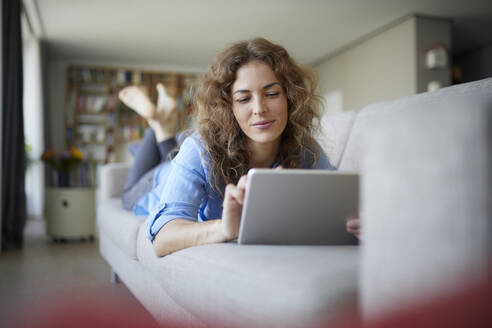 Schöne Frau, die ein digitales Tablet benutzt, während sie zu Hause auf dem Sofa liegt - RBF07956