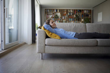 Frau mit Kopfhörern hört Musik, während sie zu Hause auf dem Sofa liegt - RBF07958