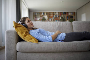 Mid adult woman wearing headphones listening music while lying on sofa at home - RBF07959