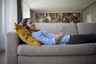 Mittlere erwachsene Frau mit Kopfhörern, die Musik hört, während sie zu Hause auf dem Sofa liegt - RBF07959