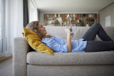 Mittlere erwachsene Frau, die ein digitales Tablet benutzt, während sie zu Hause auf dem Sofa liegt - RBF07960
