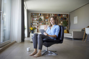 Lächelnde Frau winkt mit der Hand zu einem Videoanruf auf einem digitalen Tablet, während sie auf einem Stuhl zu Hause sitzt - RBF07965