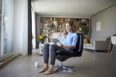 Smiling woman waving hand to video call on digital tablet while sitting on chair at home - RBF07965